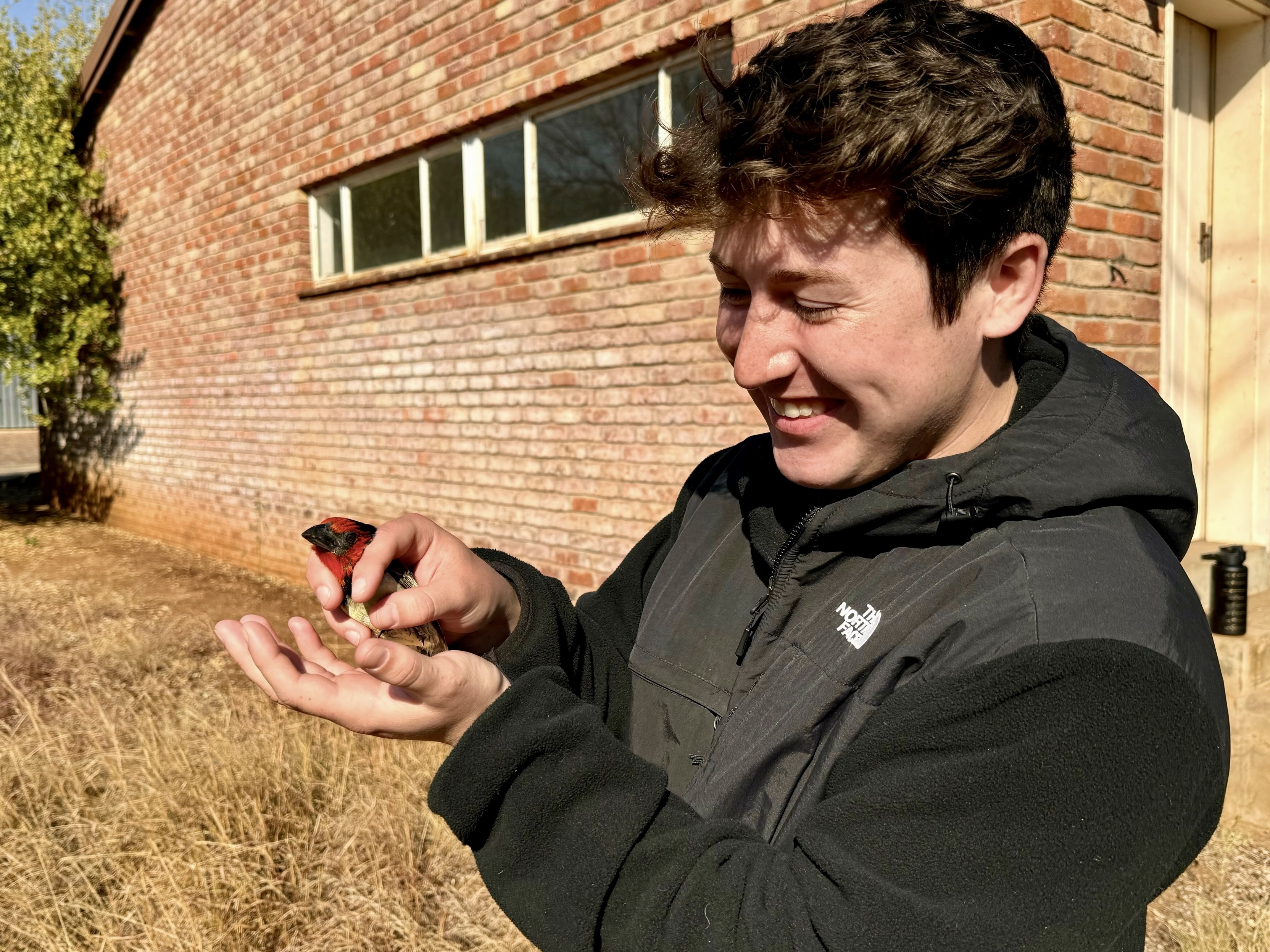 Michael is standing outside wearing a black jacket and smiling while he holds a black collared barbet.