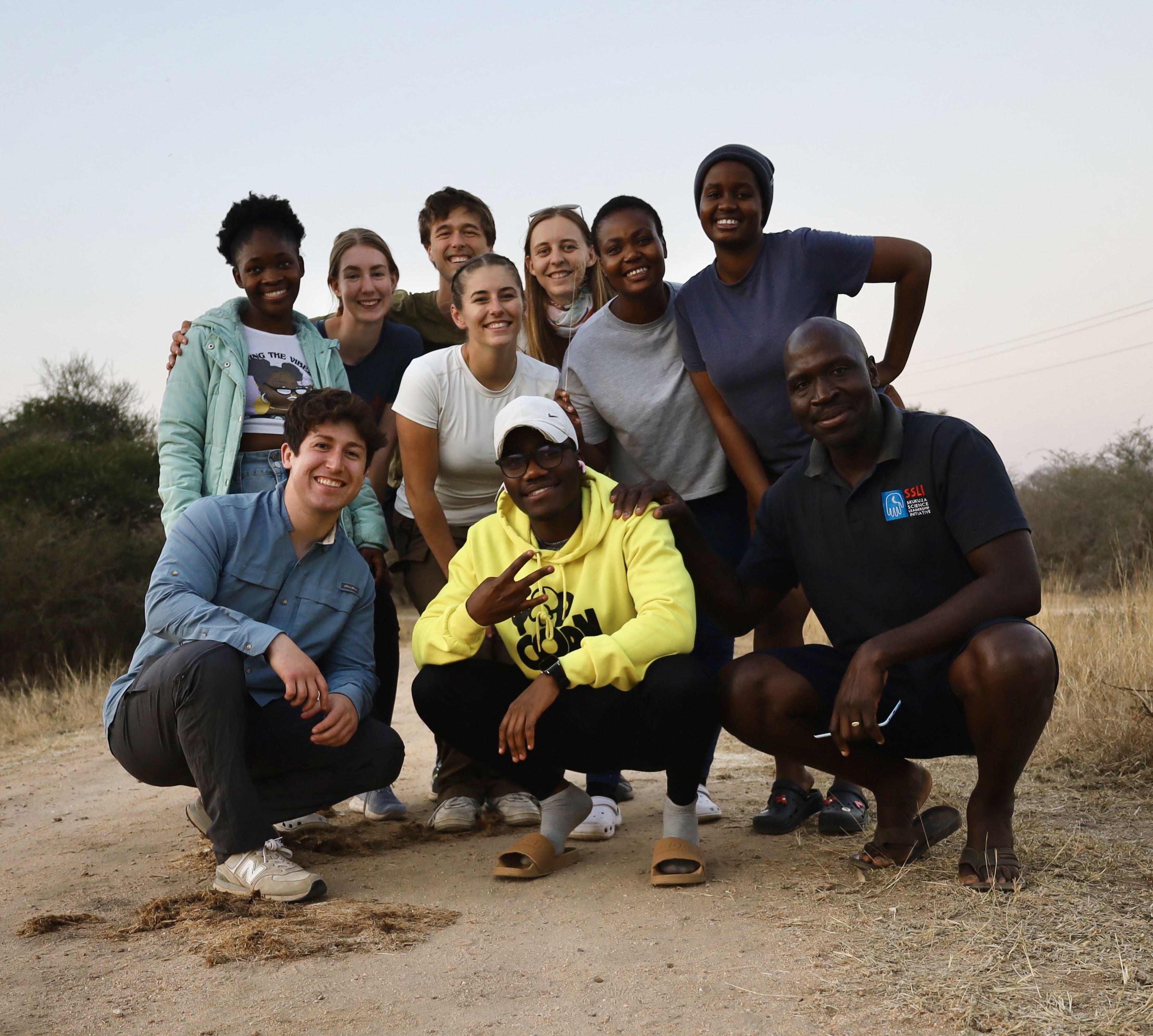 Several people squatting down on a dirt path, smiling at the camera.